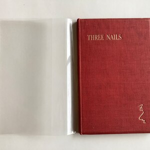 May include: A red hardback book with the title "THREE NAILS" in gold lettering. The book has a textured cover and a gold emblem in the lower right corner. A clear plastic sleeve is open to the left of the book.