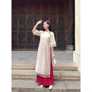 May include: A white traditional Vietnamese dress with pink trim and floral embroidery. The dress is worn with a pair of bright pink pants.