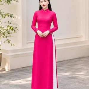 May include: A vibrant fuchsia long-sleeved tunic with a high collar and side slits, featuring delicate white pearl-like bead accents along the neckline, sleeve cuffs, and side openings, worn over loose-fitting white trousers.