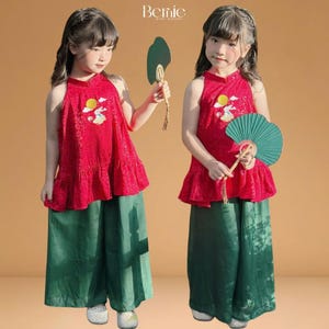 以下が含まれることがあります： お揃いの衣装を着た2人の若い女の子。赤いホルターネックのトップスとグリーンのワイドパンツ。トップスには太陽とウサギの刺繍が施されています。各女の子は緑の扇子を持っています。ブランド名「Bernie」が表示されています。