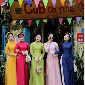 May include: Five women wearing traditional Vietnamese áo dài dresses in various colors, including yellow, pink, red, green, and blue. The women are standing in front of a shop with a sign that reads "CÚB cà phê".
