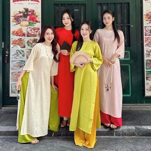 May include: Four women wearing traditional Vietnamese dresses, known as Ao Dai, in different colors: white, red, yellow, and pink. They are standing in front of a green door with a sign above it.