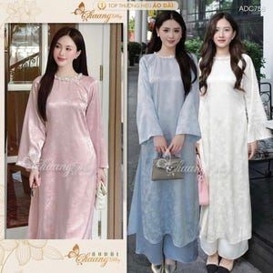 Puede incluir: Tres mujeres con vestidos largos y fluidos con un collar de perlas. Los vestidos son de color rosa, azul claro y blanco. Las mujeres están de pie frente a un edificio con una puerta de vidrio. El texto "TOP THUONG HIEU AO DAI" es visible en la parte superior de la imagen.
