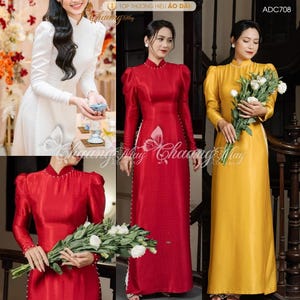 May include: Three long, elegant áo dài dresses in white, red, and yellow. The dresses feature high collars, long sleeves, and pearl accents. The red and yellow dresses are held by women holding bouquets of white flowers. The dresses are formal wear.