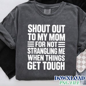 May include: A charcoal grey short-sleeved t-shirt with a distressed white graphic print that reads: SHOUT OUT TO MY MOM FOR NOT STRANGLING ME WHEN THINGS GET TOUGH. The garment features a crew neckline and a vintage-style washed fabric texture.