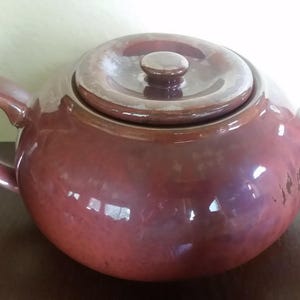 May include: A maroon ceramic teapot with a matching lid and handle. The teapot has a rounded body and a curved spout. The handle is arched, and the lid has a small knob. The teapot is sitting on a wooden surface.