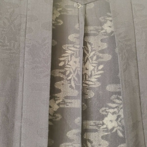 May include: Black kimono with a subtle pattern of white flowers and leaves. The kimono is closed with a single button at the top.