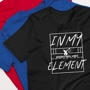 以下が含まれることがあります： 白いグラフィックで「IN MY ELEMENT」と書かれた黒いTシャツ。小さなロゴには「RACQUETBALL VIBEZ」と書かれています。Tシャツは折り畳まれて、青いTシャツと赤いTシャツの上に置かれています。