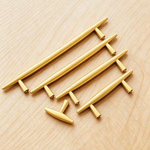 May include: A collection of gold-toned cabinet pulls and handles in various sizes. The hardware features a sleek, modern design with a cylindrical bar and T-shaped ends. The pulls are arranged on a light wood surface.