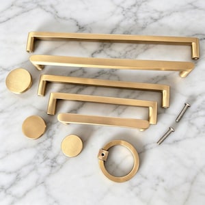 May include: A collection of brushed brass cabinet hardware including four rectangular bar pulls of varying lengths, three circular knobs, one circular ring pull, and two silver-toned metal mounting screws arranged on a white marble surface.
