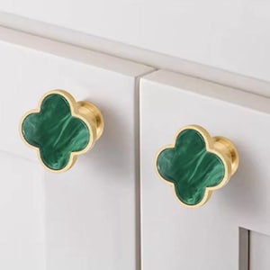 May include: Two cabinet knobs with a four-leaf clover design. The knobs have a gold-colored base and a green, marbled clover-shaped center. The knobs are mounted on a white cabinet door.