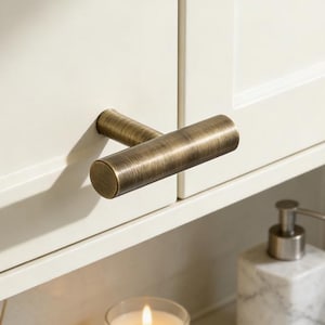 May include: A close-up of a cabinet door with a bronze-colored, cylindrical handle. The cabinet is off-white. A lit candle in a glass jar, a marble soap dispenser, and a reed diffuser are visible on the countertop.