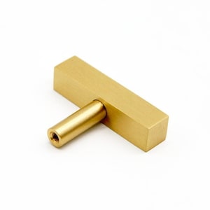 May include: A gold-toned, rectangular cabinet knob with a straight, cylindrical handle.
