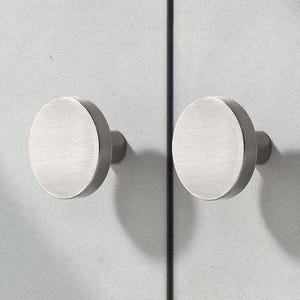 May include: Two brushed nickel cabinet knobs with a circular design. The knobs are mounted on a light gray cabinet door. The knobs have a simple, modern aesthetic.