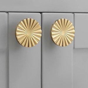 May include: Two gold-toned round cabinet knobs with a sunburst design. The knobs have a raised, textured surface.