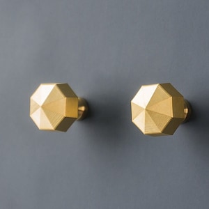 May include: Two gold-colored, geometric cabinet knobs with a faceted design. The knobs are octagonal and have a brass finish. They are mounted on a gray surface, likely a cabinet door or drawer front.