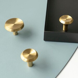 May include: Three gold-toned metal cabinet knobs with a round, slightly raised top and a short, cylindrical stem. The knobs are shown on a light blue and black surface.