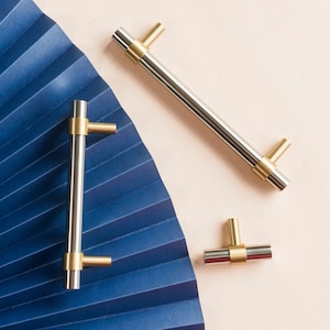 Op de afbeelding: Drie kastknoppen met een geborsteld nikkel afwerking en gouden accenten. De knoppen bevinden zich op een lichtroze achtergrond.