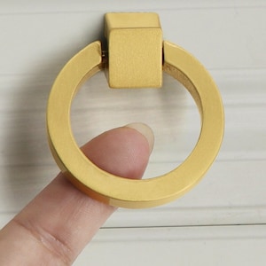 May include: Gold ring pull cabinet hardware with a square top. The ring pull is made of metal and has a smooth, brushed finish.