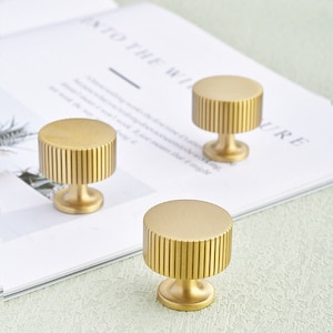 May include: Three gold cabinet knobs. Each knob has a cylindrical body with vertical grooves and a rounded top, mounted on a short, pedestal base. They are displayed on a white surface with text.