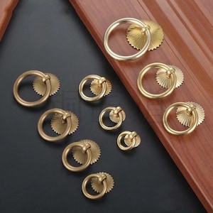 May include: A collection of brass-colored ring pulls for cabinets or drawers. Each pull features a circular handle attached to a decorative, sunburst-shaped backplate. The hardware is arranged on a dark surface with a wooden accent.