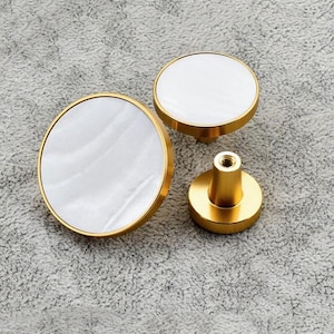 May include: Three round cabinet knobs with a white marble-like surface and a gold metal rim. The knobs are different sizes.