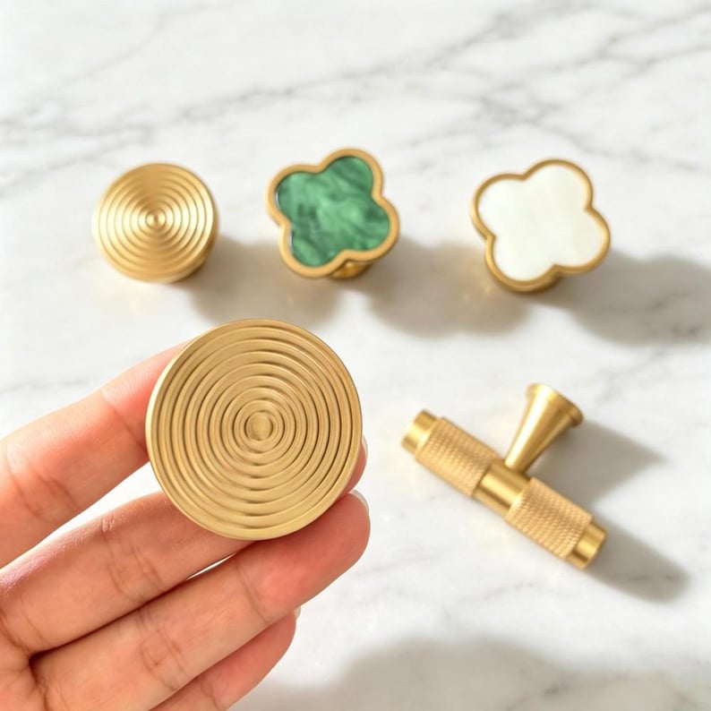 Peut inclure: Une collection de quincaillerie d'ameublement comprenant deux boutons dor&eacute;s circulaires &agrave; motifs d'anneaux concentriques, un bouton en forme de tr&egrave;fle avec incrustation marbr&eacute;e verte, un bouton en forme de tr&egrave;fle blanc et une poign&eacute;e en T dor&eacute;e.