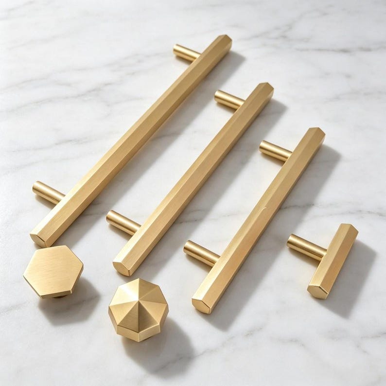 May include: A collection of brushed gold cabinet hardware including three long hexagonal bar pulls and two matching knobs. The hardware features a metallic finish with clean geometric lines and faceted edges, showcasing a uniform brass-toned appearance.