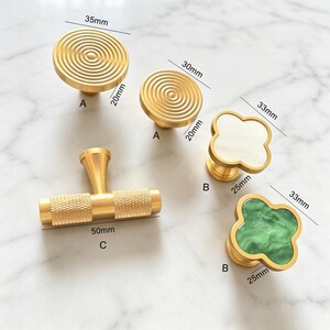 Peut inclure: Une collection de boutons et poign&eacute;es de placard dor&eacute;s. Deux boutons ronds &agrave; cercles concentriques mesurent 35mm et 30mm de diam&egrave;tre. Deux boutons en forme de tr&egrave;fle avec incrustations blanches et vertes mesurent 33mm. Une poign&eacute;e en T molet&eacute;e mesure 50mm.