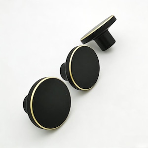 Puede incluir: Tres pomos de armario negros con un borde dorado. Los pomos tienen un dise&ntilde;o redondo y plano y est&aacute;n colocados sobre un fondo blanco. Los pomos son probablemente para muebles o armarios.