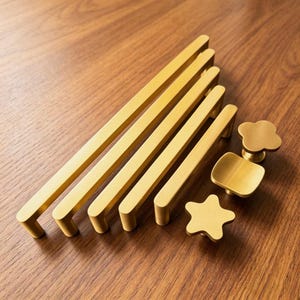 May include: A collection of brushed gold cabinet hardware including four long rectangular bar pulls of varying lengths and three decorative knobs shaped like a four-leaf clover, a rounded square, and a five-pointed star with a matte finish.