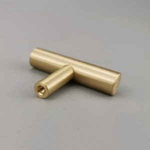 May include: A gold-toned T-shaped cabinet knob with a smooth, polished finish.