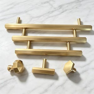 May include: A collection of brushed gold hardware including three horizontal bar pulls of varying lengths, a faceted hexagonal knob, a small rectangular T-bar pull, and a single hexagonal flat-faced knob, all featuring a smooth metallic finish.
