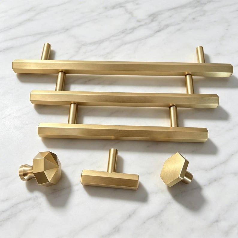 May include: A collection of brushed gold hardware including three horizontal bar pulls of varying lengths, a faceted hexagonal knob, a small rectangular T-bar pull, and a single hexagonal flat-faced knob, all featuring a smooth metallic finish.