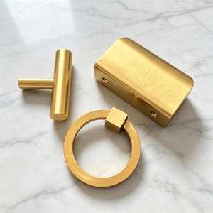 May include: A set of three gold-coloured metal hardware pieces consisting of a T-shaped bar handle, a rectangular block handle with two visible screw holes on the side, and a circular ring pull attached to a small square mounting base.