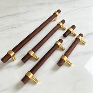 May include: A collection of five cabinet pulls featuring cylindrical dark brown wood bars accented with brushed gold metal mounting posts. The set includes one small knob and four elongated handles of varying lengths, all with a smooth finish.