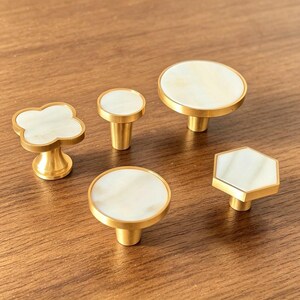 Peut inclure: Un ensemble de cinq boutons de placard avec des bases en métal doré et des incrustations blanches lisses, de formes circulaire, trèfle et hexagonale. Le lot comprend un bouton en forme de trèfle, un petit bouton rond, deux grands boutons ronds et un bouton hexagonal.
