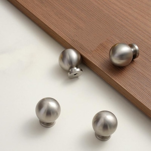 May include: Four brushed nickel cabinet knobs with a spherical design. The knobs are arranged on a white surface with a brown wooden edge in the background. The knobs have a simple, modern aesthetic.
