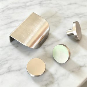 Peut inclure: Un ensemble de quincaillerie d'armoire comprenant une grande poignée profilée en métal argenté brossé avec un profil incurvé et deux petits boutons circulaires en métal argenté brossé à face plate, disposés pour montrer leur finition.