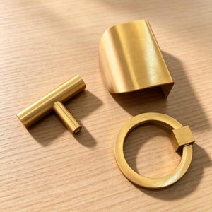 Peut inclure: Trois pièces de quincaillerie d'armoire en or brossé comprenant une poignée en T, une poignée de bord incurvée et une poignée annulaire circulaire avec une base de montage carrée. Chaque pièce présente une finition métallique lisse et dorée.
