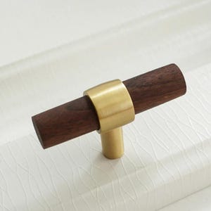 Peut inclure: Poignée de meuble avec une poignée en bois brun foncé et une base en métal de couleur laiton. La poignée est cylindrique et la base est un anneau qui relie la poignée à une courte tige cylindrique.