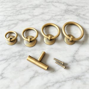 May include: A set of four brass ring pulls with circular, sunburst-patterned backplates in graduated sizes, accompanied by one T-shaped bar handle and two matching metal mounting screws, all featuring a brushed, matte gold-toned finish.