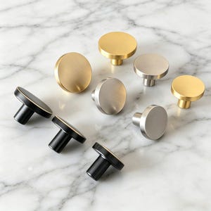 May include: A collection of eight circular cabinet knobs arranged in a group. The set includes three matte black knobs, three brushed gold knobs, and two brushed silver knobs. Each knob features a flat, round face and a cylindrical base.