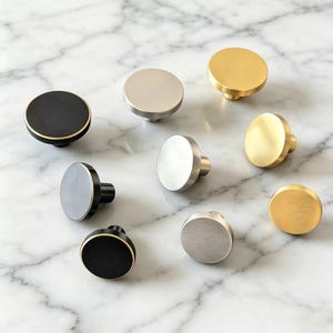 May include: A collection of nine circular cabinet knobs arranged in a group. The knobs feature three distinct finishes: matte black with gold edges, brushed silver, and brushed gold. Each knob has a flat, round face and a cylindrical base.