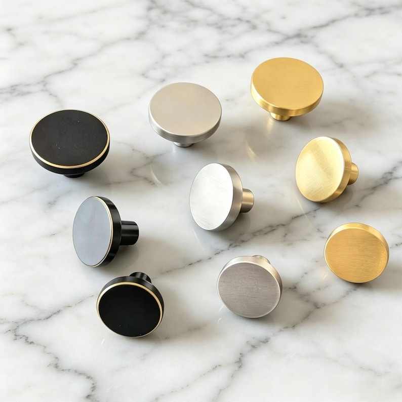 May include: A collection of nine circular cabinet knobs arranged in a group. The knobs feature three distinct finishes: matte black with gold edges, brushed silver, and brushed gold. Each knob has a flat, round face and a cylindrical base.