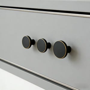 Puede incluir: Tres tiradores de armario negros con bordes dorados est&aacute;n montados en un caj&oacute;n gris claro. Los tiradores son redondos y espaciados uniformemente, con un dise&ntilde;o elegante y moderno. El caj&oacute;n tiene un borde plateado.