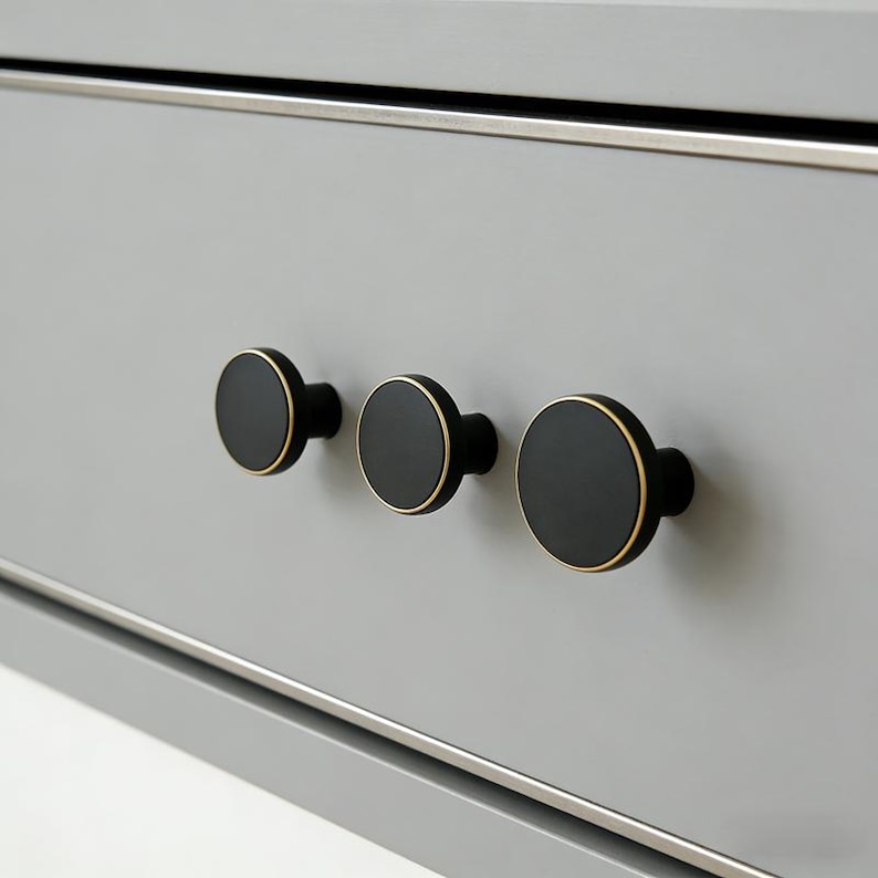 Puede incluir: Tres tiradores de armario negros con bordes dorados est&aacute;n montados en un caj&oacute;n gris claro. Los tiradores son redondos y espaciados uniformemente, con un dise&ntilde;o elegante y moderno. El caj&oacute;n tiene un borde plateado.