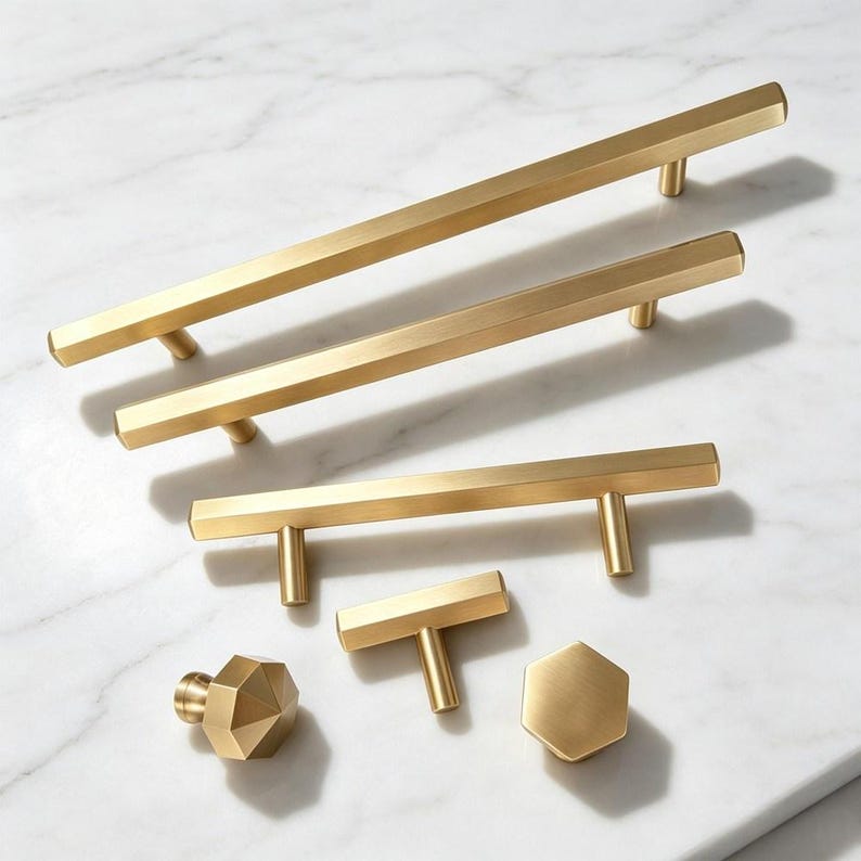 May include: A collection of brushed brass cabinet hardware including three long bar pulls of varying lengths, one T-shaped handle, one hexagonal knob, and one faceted knob. Each piece features a smooth, metallic gold finish and geometric shapes.