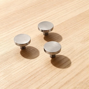 May include: Three brushed nickel cabinet knobs with a circular design. The knobs are set against a light wood grain surface, casting shadows. These modern knobs are suitable for drawers and cabinets.