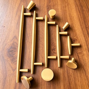 May include: A collection of brushed gold cabinet hardware including four long cylindrical bar pulls with two mounting points each, one T-shaped handle, and three round knobs with a flat top surface and a tapered base, all arranged on a wooden surface.